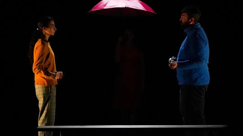 Two actors stand in the dark facing each other under a red umbrella.