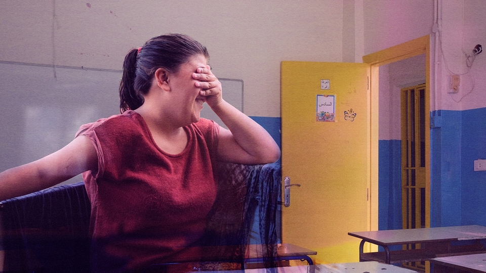 A composite of two images: a girl covering her eyes and looking away with a shy smile on her face in the foreground, with a classroom in the background.