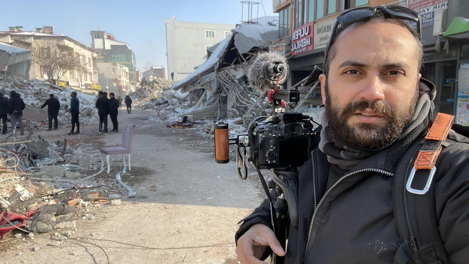 A self-portrait of journalist Issam Abdallah in the foreground, carrying his DSLR camera and microphone, to the background of destroyed buildings. Clear winter day.
