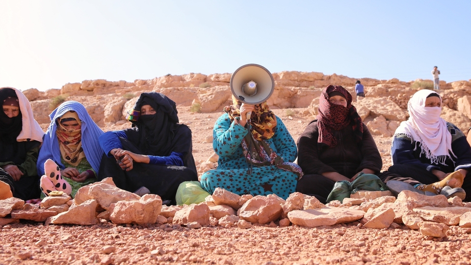 A group of six Amazigh women sit on the ground shoulder-to-shoulder. They all have their hair and faces partially covered; one of them is talking into a megaphone.