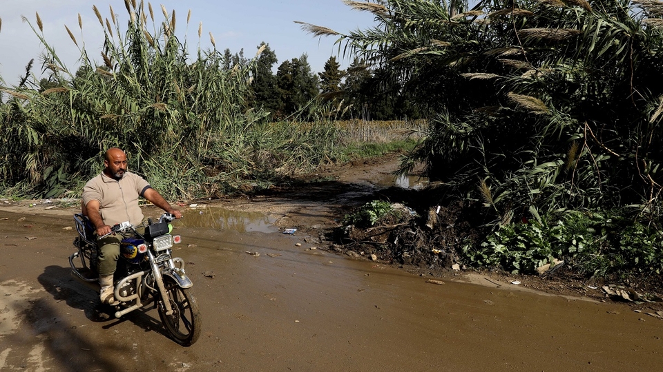 Khaled Al Masri arriving on his motorcycle on a road flooded with sewage, with bamboo sticks emerging from the side of the road in Akkar.